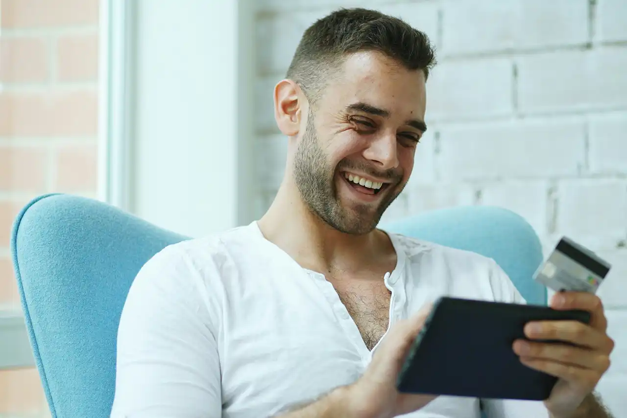 Un homme, muni d'une carte de crédit et d'une tablette, est assis sur un canapé.